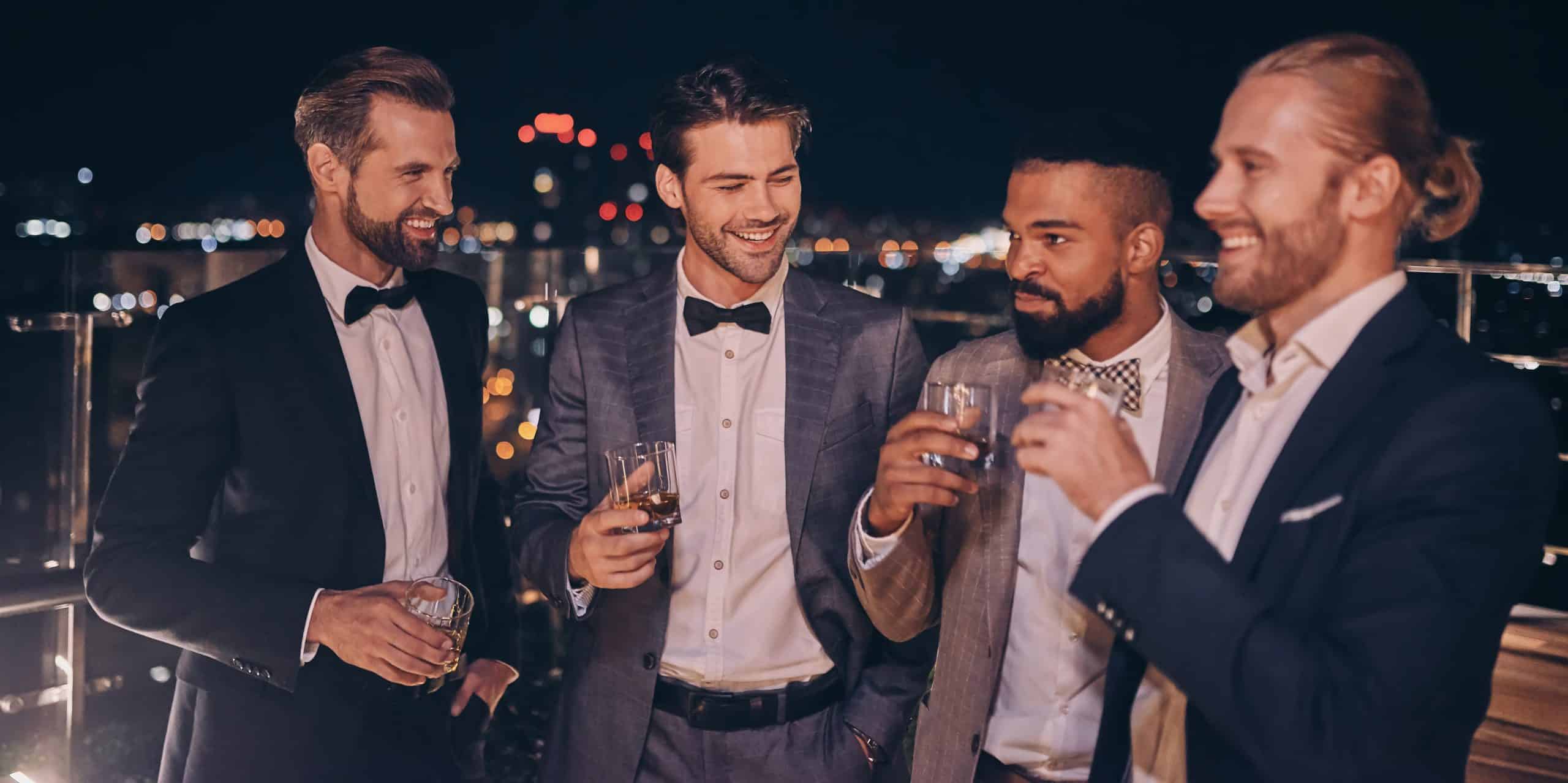 Group of handsome young men in suits and bowties drinking whiskey and smiling while spending time on outdoor party