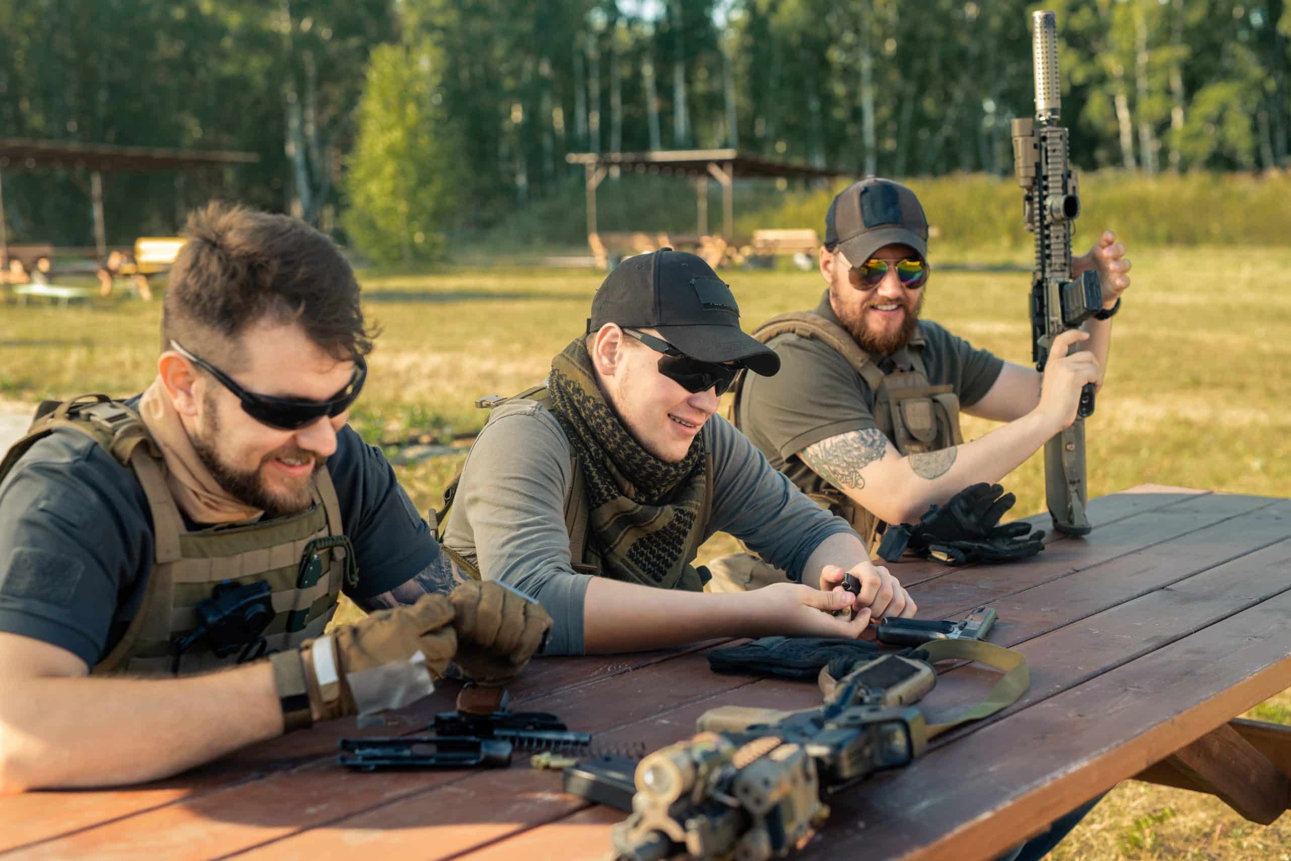 Positive young men in body armors sitting at wooden table and chatting while reloading guns